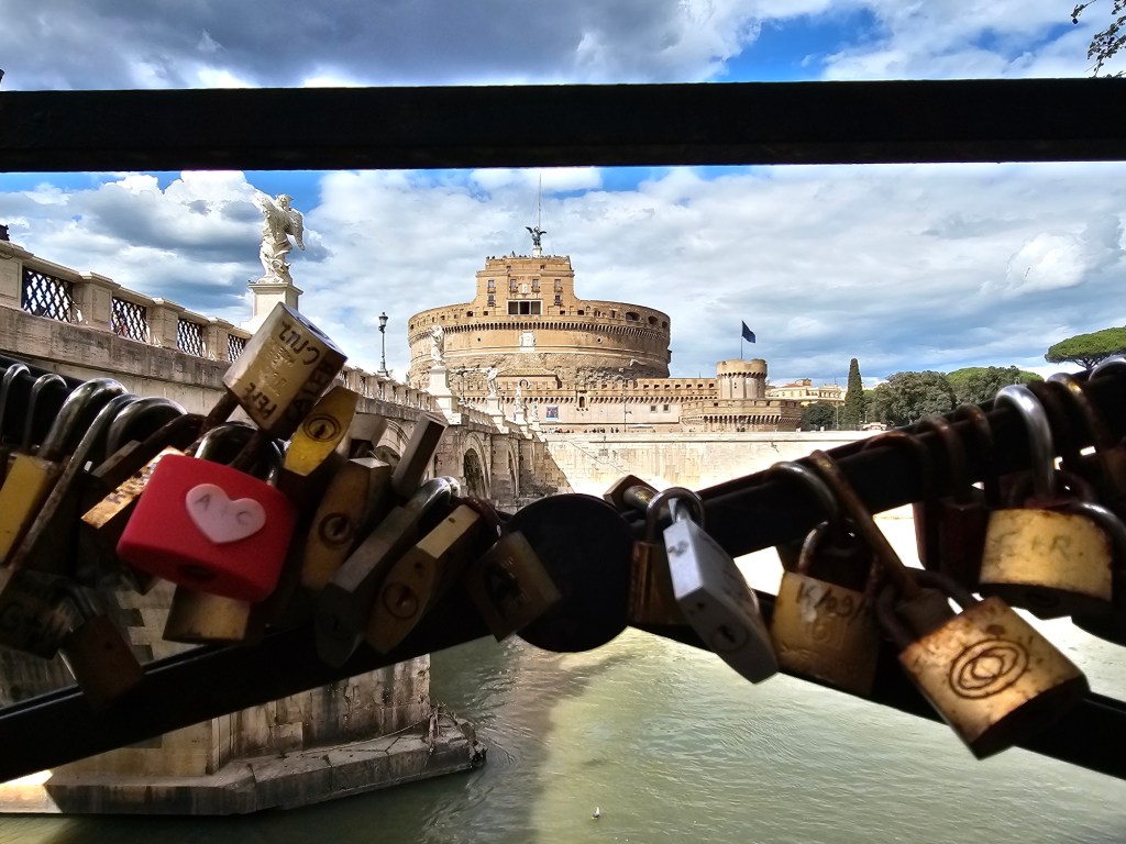 Vista do Castelo Sant'Angelo em Roma, com cadeados pendurados em uma grade, sobre o rio, sob um céu nublado.