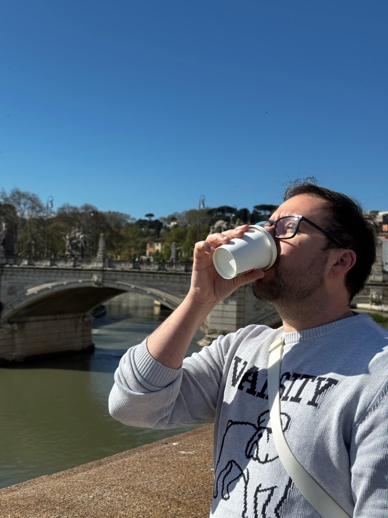 Uma pessoa bebendo café em um copo descartável, com um fundo de um rio e uma ponte em Roma sob um céu azul claro.